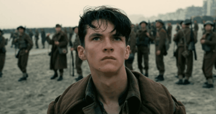 Tommy, a young British soldier, is standing on a beach looking above the camera into the ske. Beyond him are other soldiers looking up.